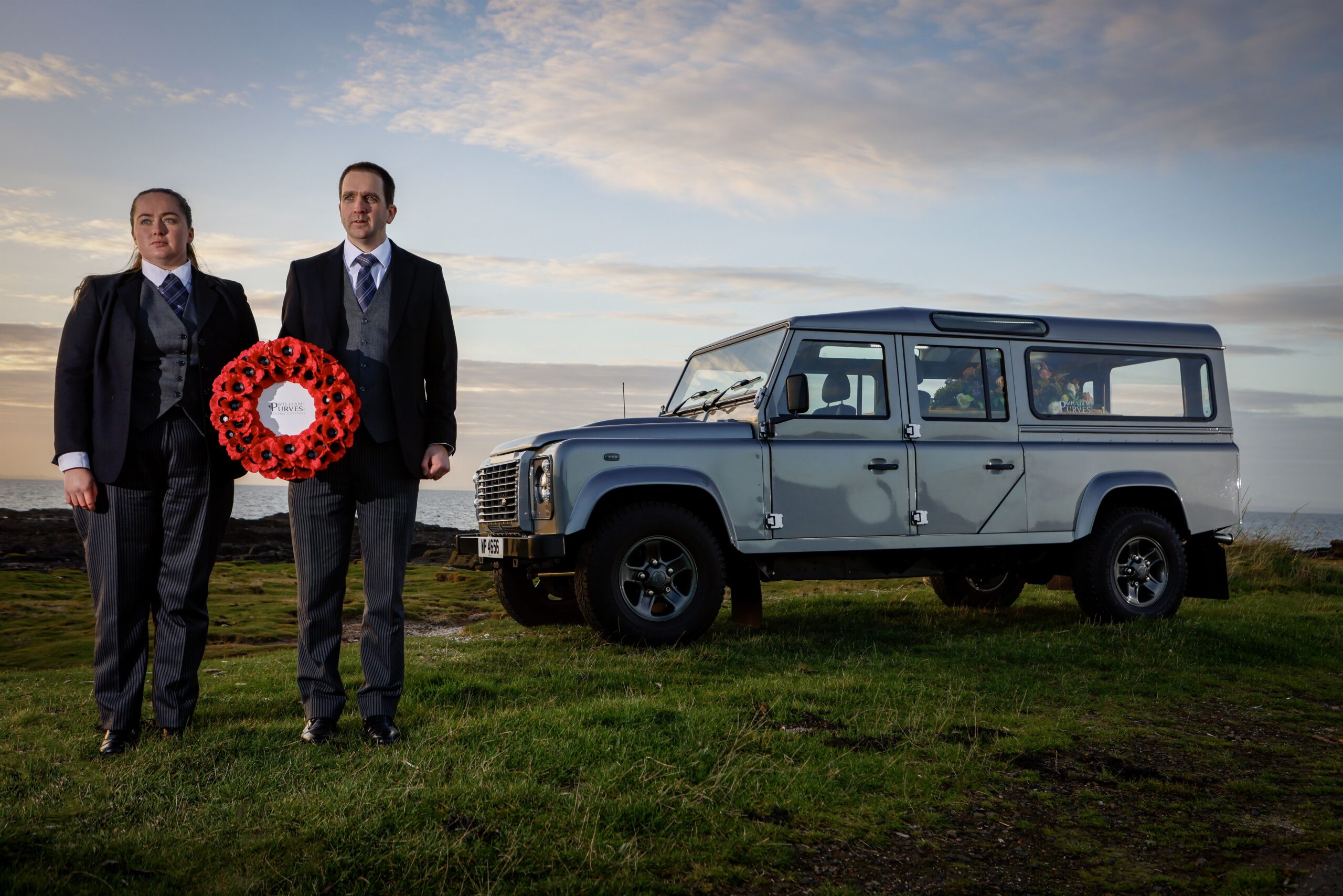 Two funeral directors dressed in formal dark suits stand on grassy terrain holding a large red poppy wreath with a white centre that reads “William Purves.” Beside them is the Land Rover Defender Hearse parked on the grass, with a scenic backdrop of cloudy sky and distant horizon.