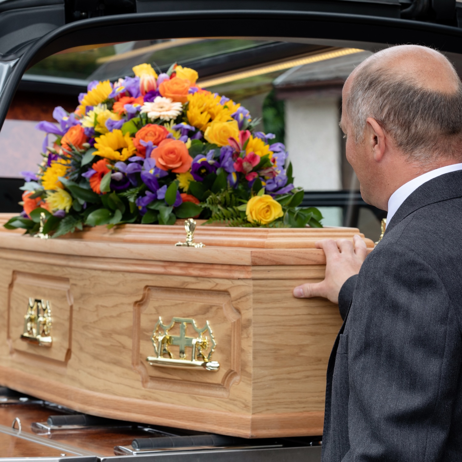 A man touching a coffin with flowers on top in the back of a funeral car