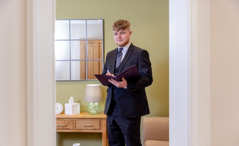 A funeral director stands with a booklet in his hands, looking at the camera